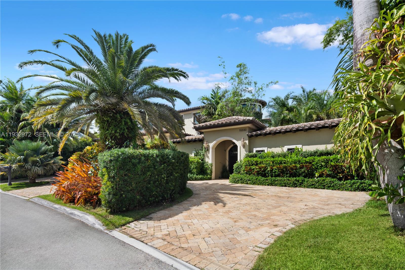 A paved driveway leads to the arched entrance of a house surrounded by lush landscaping — 10330 W Broadview Dr, Bay Harbor Islands FL 33154, BAY HARBOR ISLANDS, FL