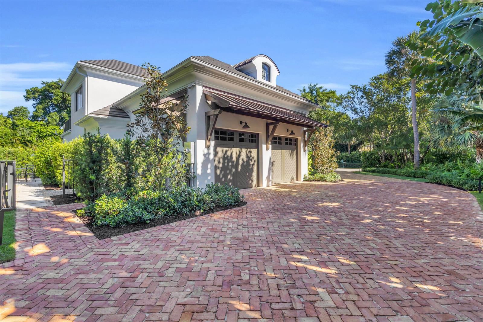 A white two-car garage with a metal awning and brick driveway sits amidst lush landscaping — 44 S Beach Road, Jupiter Island FL 33455, JUPITER ISLAND, FL