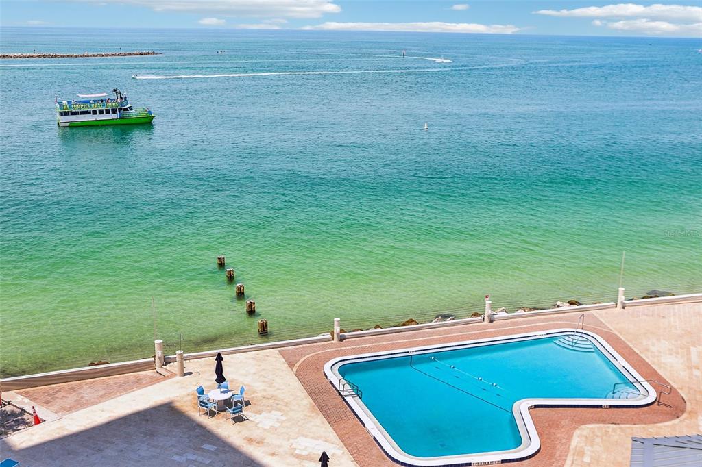 An aerial view shows a swimming pool on a patio overlooking the ocean and boat — 450 S GULFVIEW BOULEVARD UNIT 703, CLEARWATER, FL