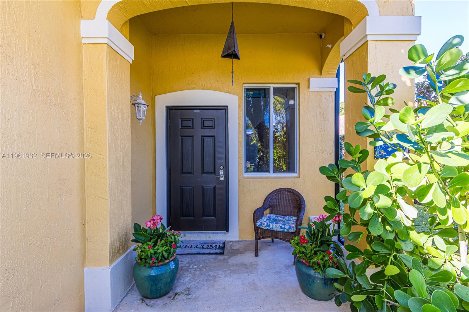 A yellow stucco entryway features a dark front door, a wicker chair, and potted plants — 1280 Majesty Ter, Weston FL 33327, WESTON, FL