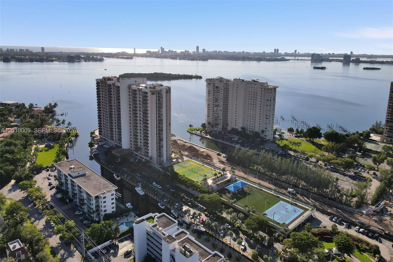 An aerial view shows high-rise apartment buildings, a tennis court, and a basketball court near water — 1800 NE 114th St # 1502, Miami FL 33181, MIAMI, FL