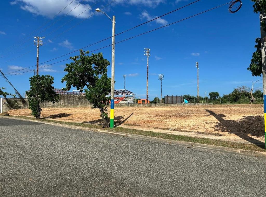 A vacant, cleared dirt lot sits next to a paved road with stadium lights behind — N BO. PUEBLO, HATILLO, PR