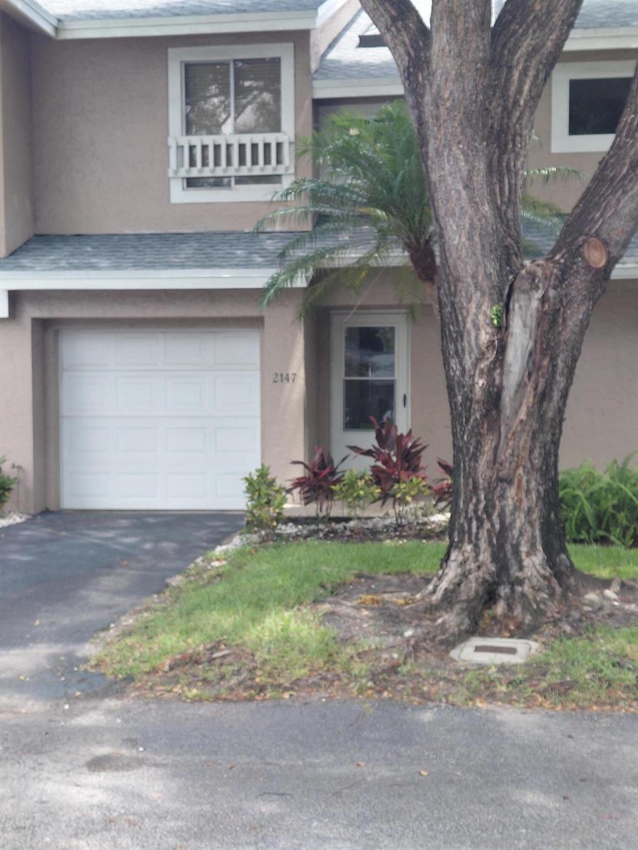 A two-story townhouse exterior with a white garage door, a front entrance, and a large tree — 2147 Discovery Circle W, Deerfield Beach FL 33442, DEERFIELD BEACH, FL