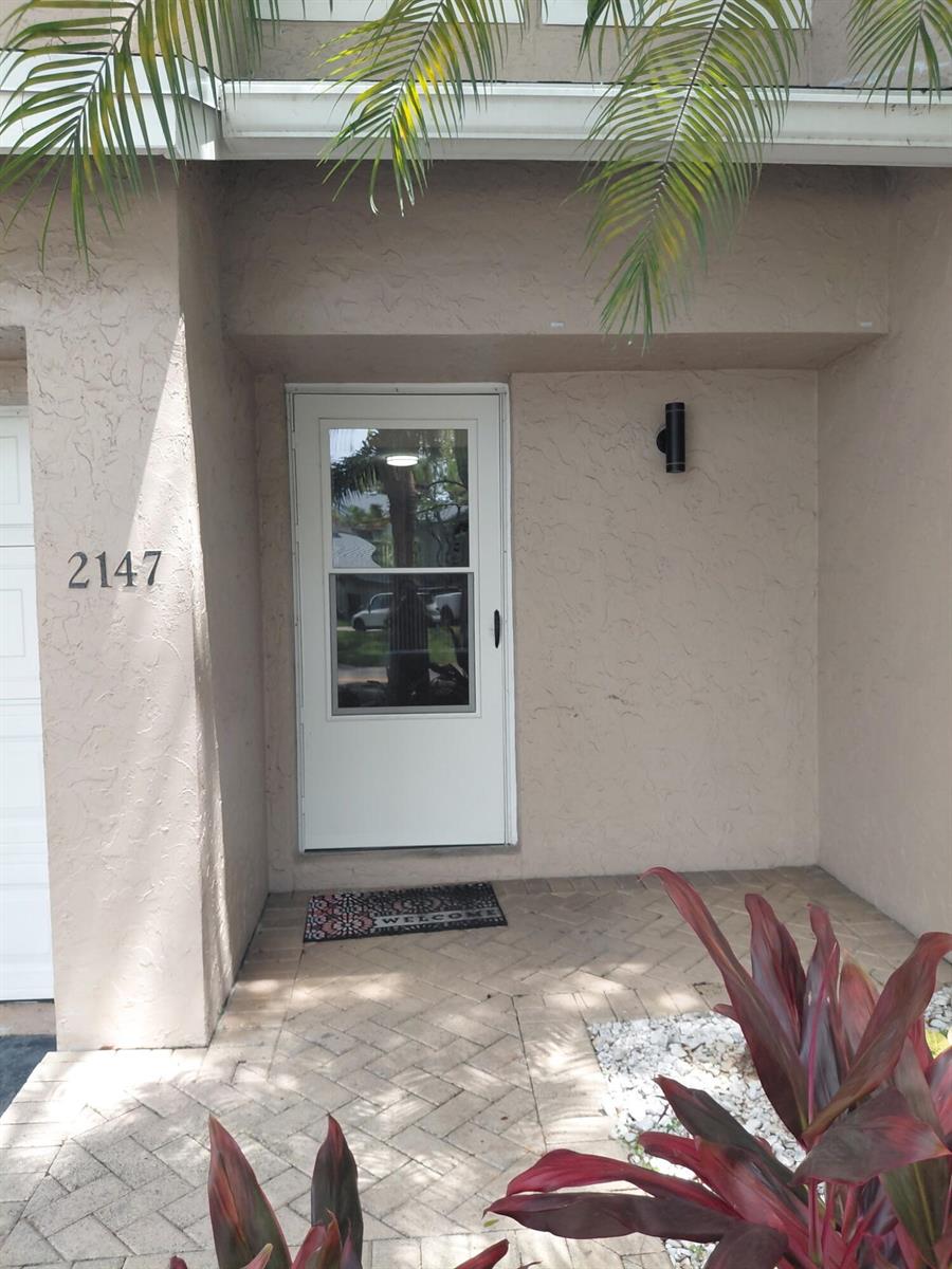 A beige exterior entryway features a white storm door, a welcome mat, and brick pavers — 2147 Discovery Circle W, Deerfield Beach FL 33442, DEERFIELD BEACH, FL