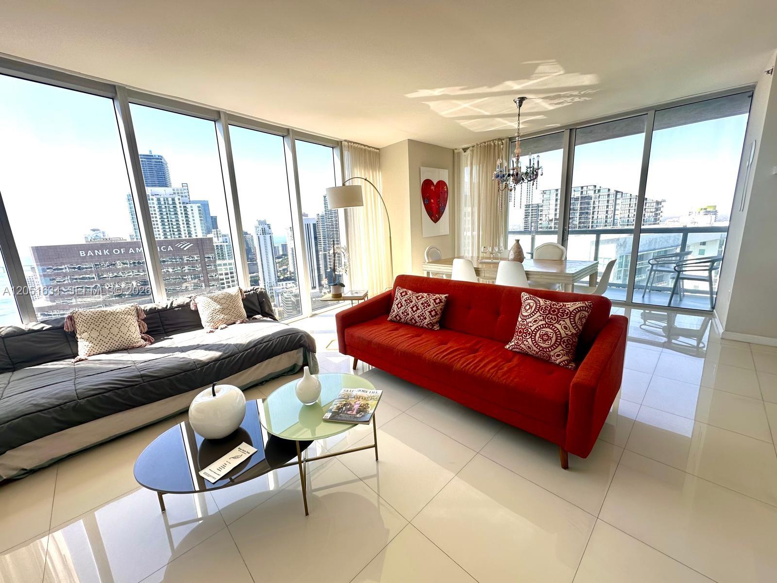 A living room with a red sofa, a grey daybed, and floor-to-ceiling windows overlooking a city — 485 Brickell Ave # 4408, Miami FL 33131, MIAMI, FL