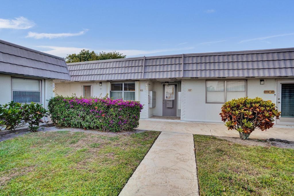 A single-story building exterior with a walkway, a purple flowering hedge, and unit numbers — 108 Valencia Boulevard # E108, Delray Beach FL 33446, DELRAY BEACH, FL