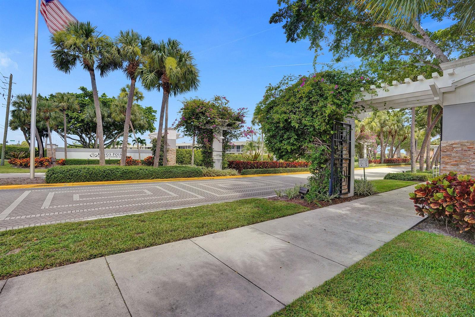 A paved entrance road leads to a gated community with palm trees and landscaping — 108 Valencia Boulevard # E108, Delray Beach FL 33446, DELRAY BEACH, FL