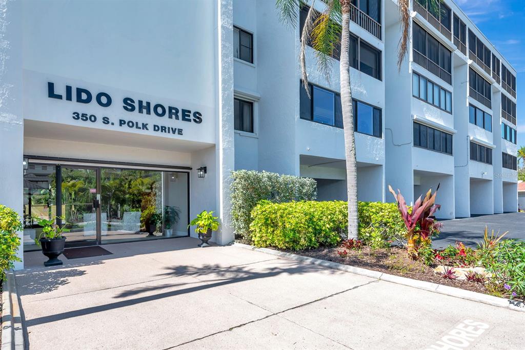 The exterior of a multi-story white apartment building features a glass entrance and landscaping — 350 S POLK DRIVE UNIT 302, SARASOTA, FL