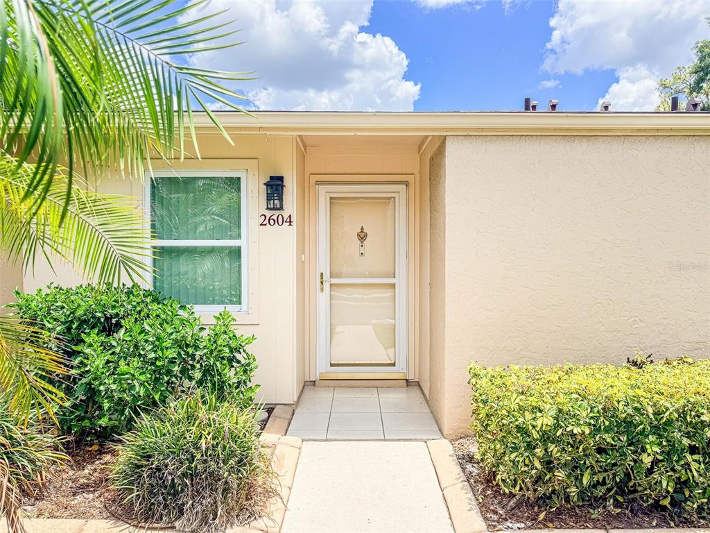 The exterior of a house features a beige stucco wall, a front door, and landscaping — 2604 WOODGATE LANE UNIT J6, SARASOTA, FL