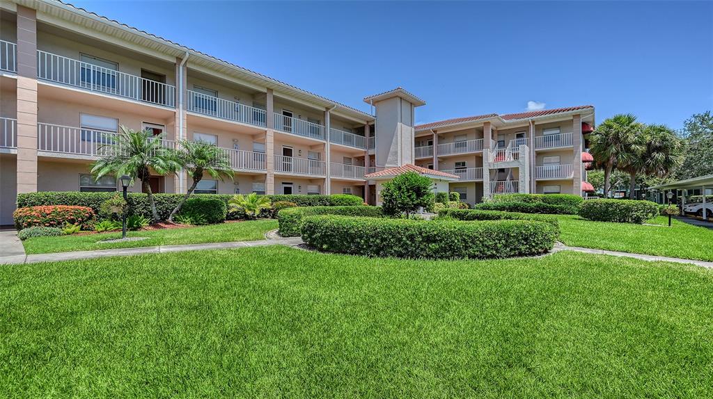 A multi-story apartment building with balconies and manicured landscaping under a clear blue sky — 6505 STONE RIVER ROAD UNIT 209, BRADENTON, FL