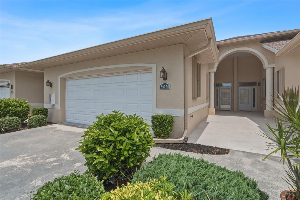 A beige house exterior features a white garage door, a covered entryway, and landscaped bushes — 24120 GREEN HERON DRIVE UNIT 6, PUNTA GORDA, FL