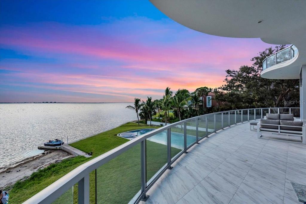 A balcony with a glass railing overlooks a swimming pool, lawn, and a large body of water — 4035 BAY SHORE ROAD, SARASOTA, FL