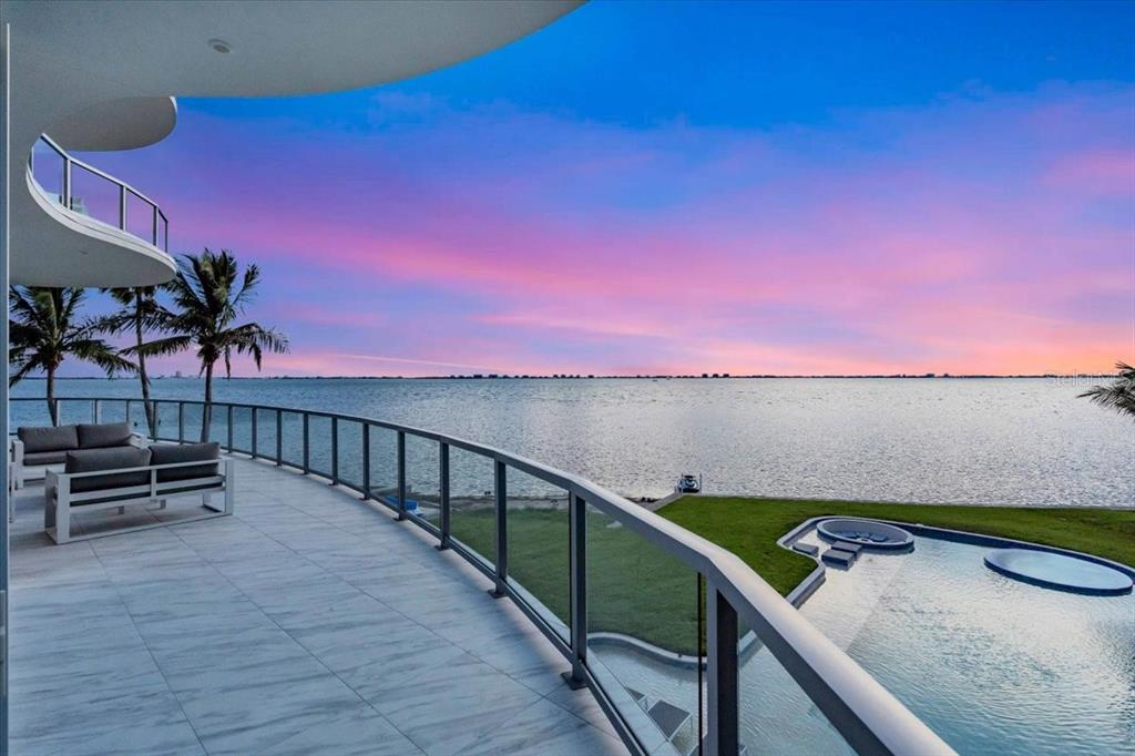 A curved balcony overlooks a swimming pool, lawn, and a body of water at sunset — 4035 BAY SHORE ROAD, SARASOTA, FL