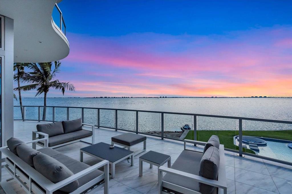 A modern outdoor balcony features gray lounge furniture overlooking a sunset view of the water — 4035 BAY SHORE ROAD, SARASOTA, FL