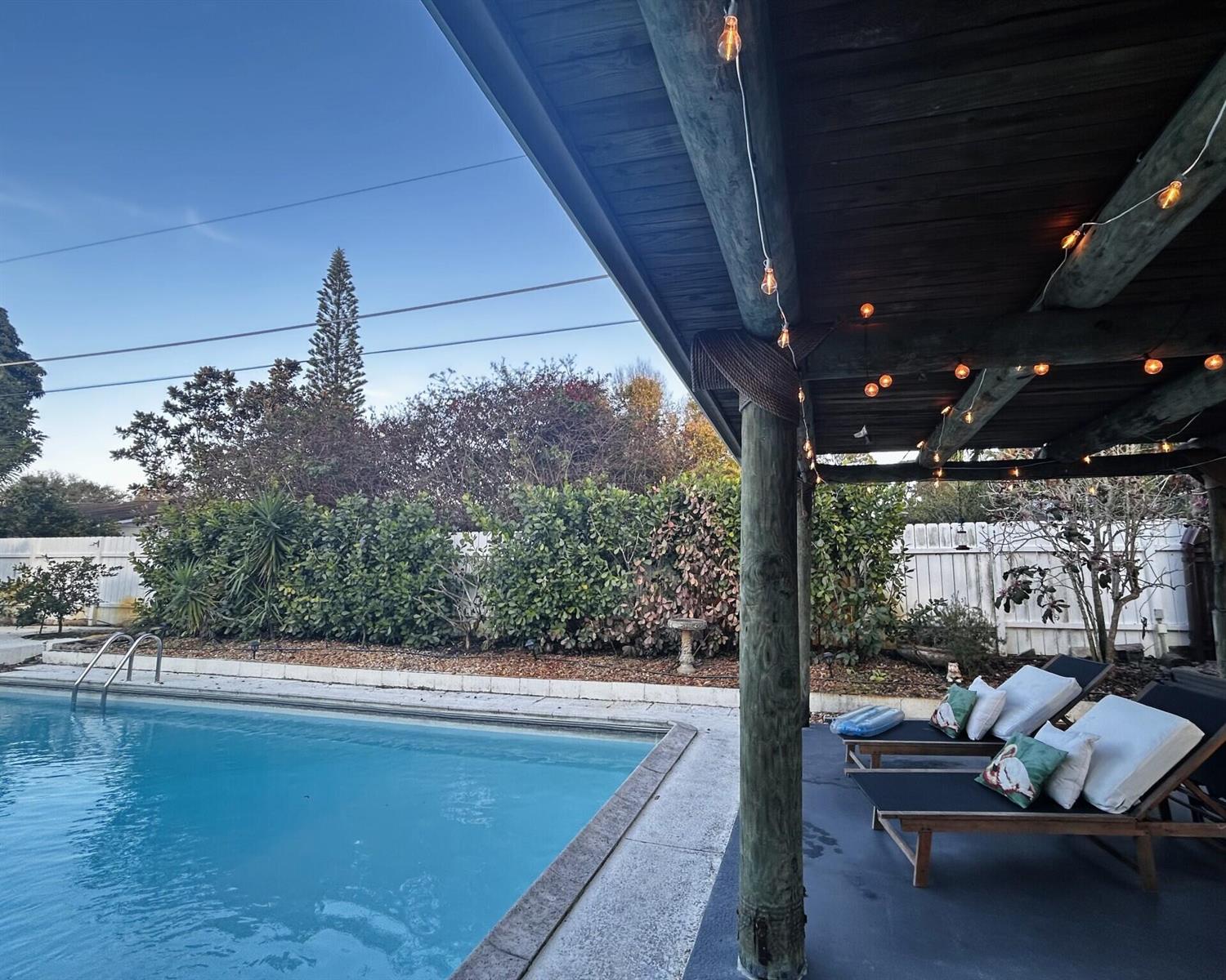 A swimming pool sits beside a covered patio featuring lounge chairs and string lights — 1301 SE Sandpiper Lane, Stuart FL 34996, STUART, FL