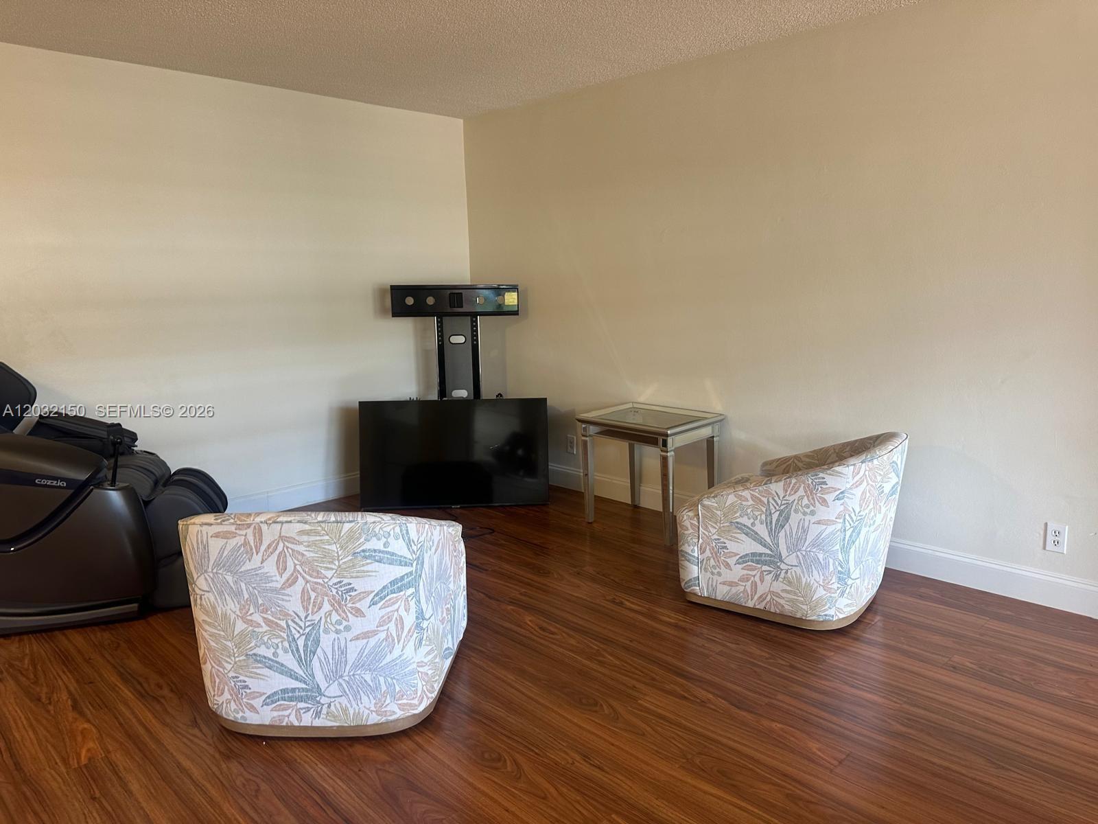 A living room features wood-look flooring, two patterned armchairs, a television, and a massage chair — 160 NE 8th Ave # 25A, Hallandale Beach FL 33009, HALLANDALE BEACH, FL