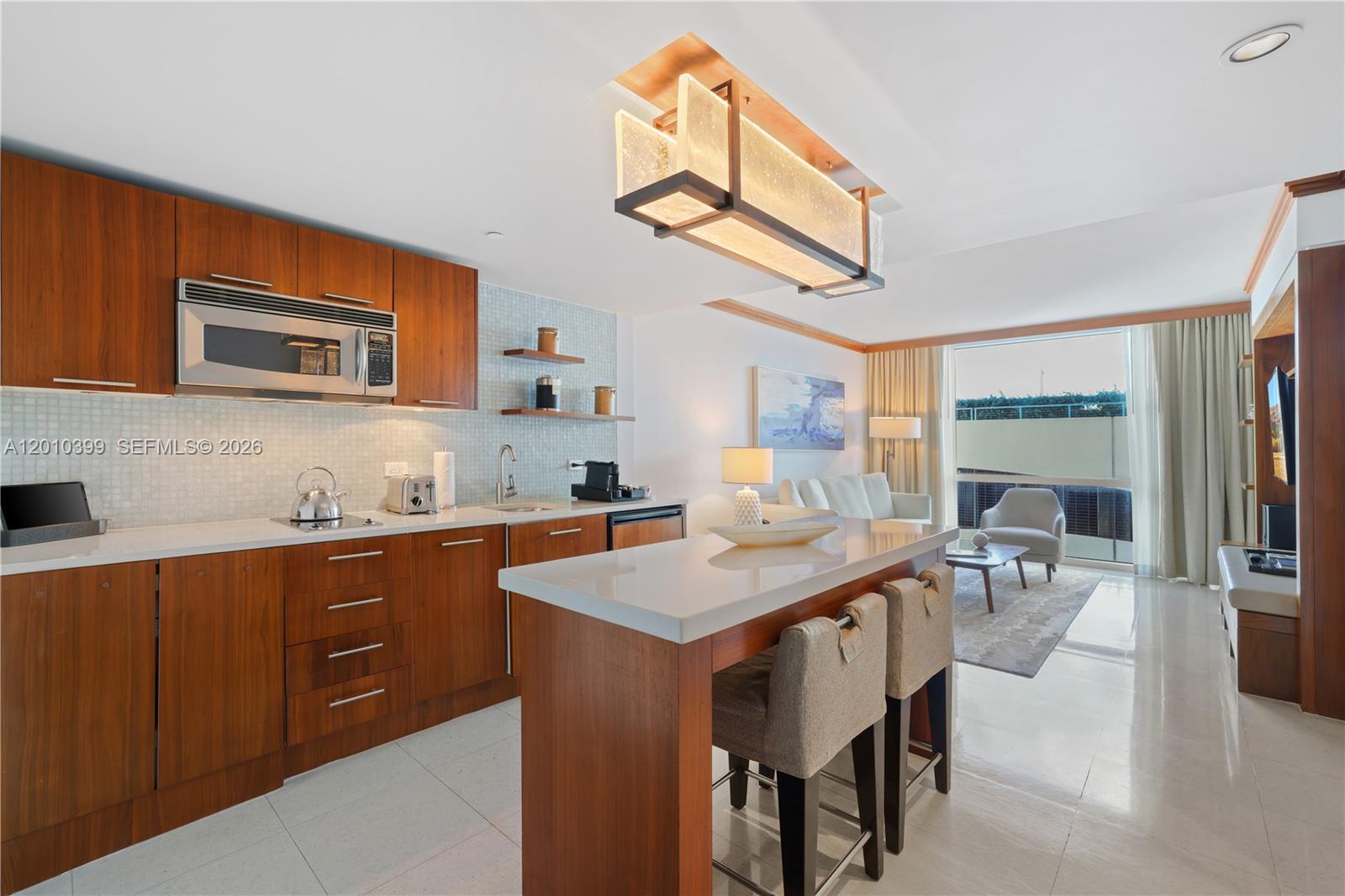 A modern kitchen with wood cabinetry and a breakfast bar opens into a living area — 6801 Collins Ave # 503, Miami Beach FL 33141, MIAMI BEACH, FL