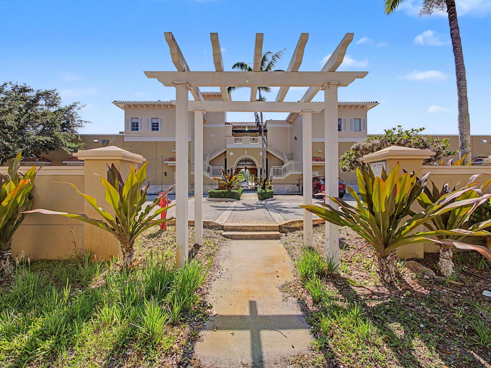 A white wooden pergola stands over a concrete walkway leading to a multi-story building entrance — 118 Black Ironwood Road # 107, Jupiter FL 33458, JUPITER, FL