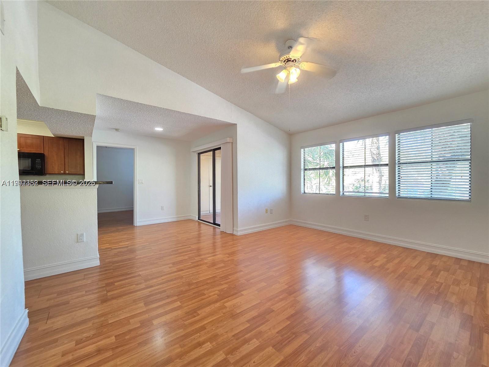An empty living room features wood-look flooring, a vaulted ceiling with a fan, and large windows — 15650 SW 105th Ln # 502, Miami FL 33196, MIAMI, FL