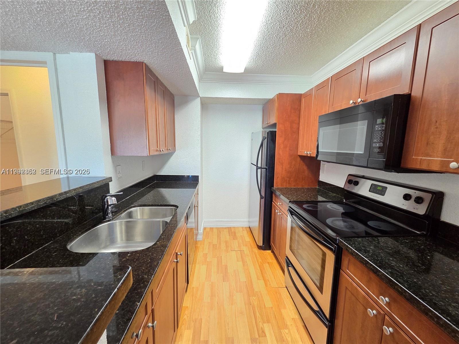 A galley kitchen features wood cabinets, black countertops, stainless steel appliances, and light wood flooring — 15650 SW 105th Ln # 502, Miami FL 33196, MIAMI, FL