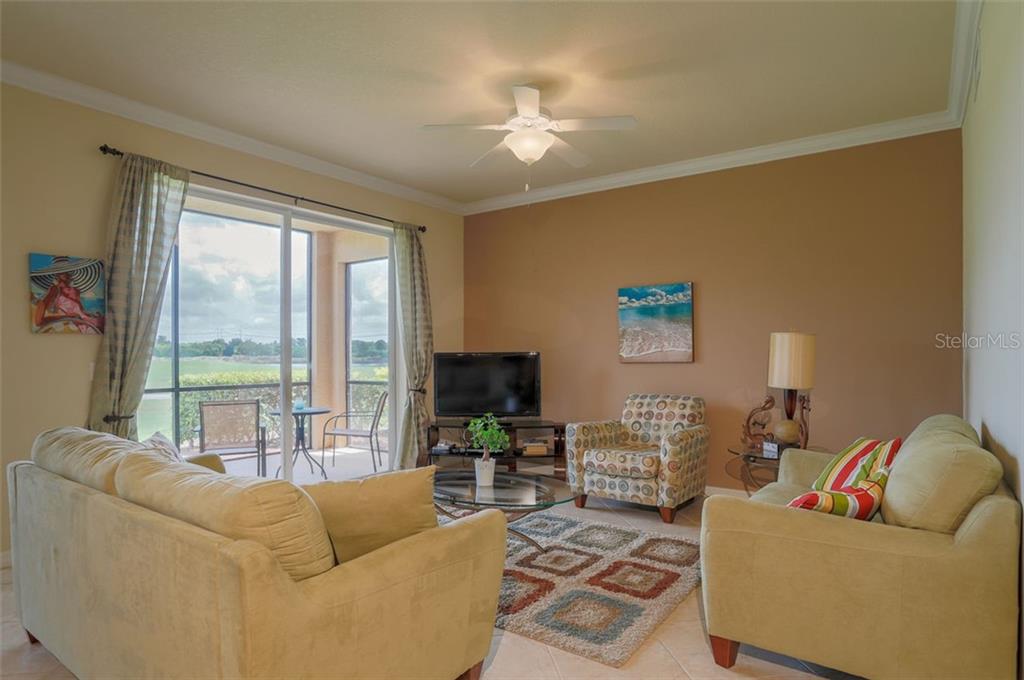 A living room features two beige sofas, a patterned armchair, a television, and sliding glass doors — 8105 GRAND ESTUARY TRAIL UNIT 106, BRADENTON, FL