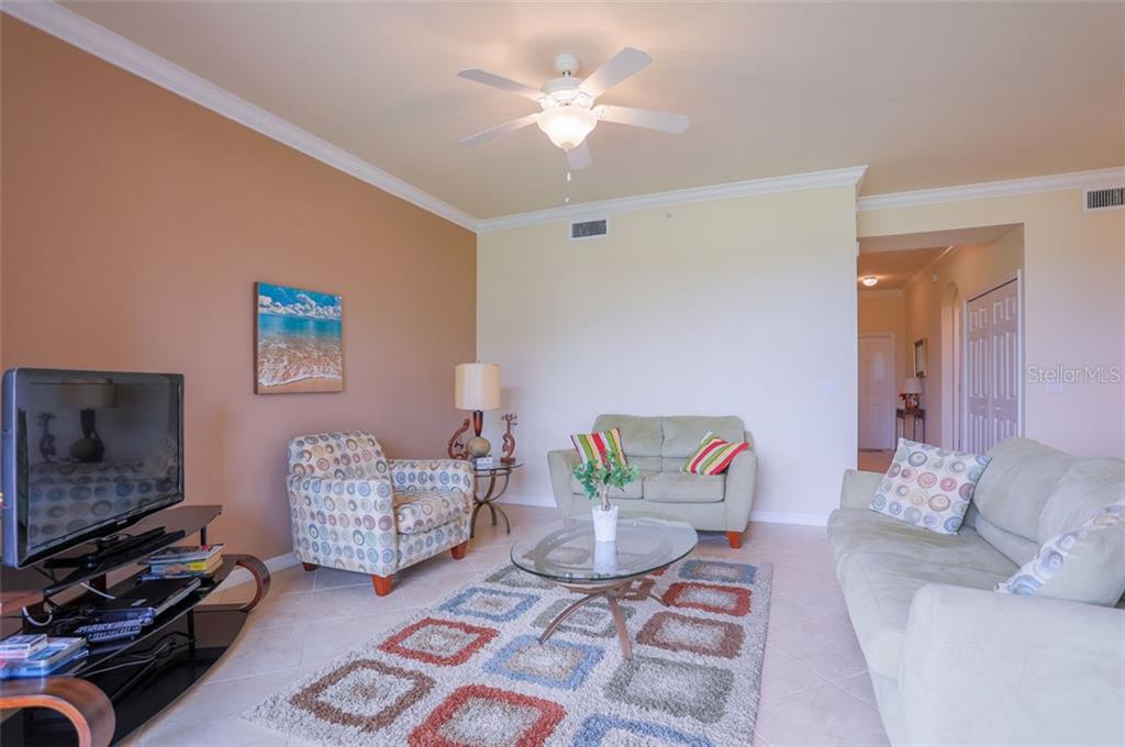 A living room features a beige sofa, patterned armchair, glass coffee table, and a ceiling fan — 8105 GRAND ESTUARY TRAIL UNIT 106, BRADENTON, FL