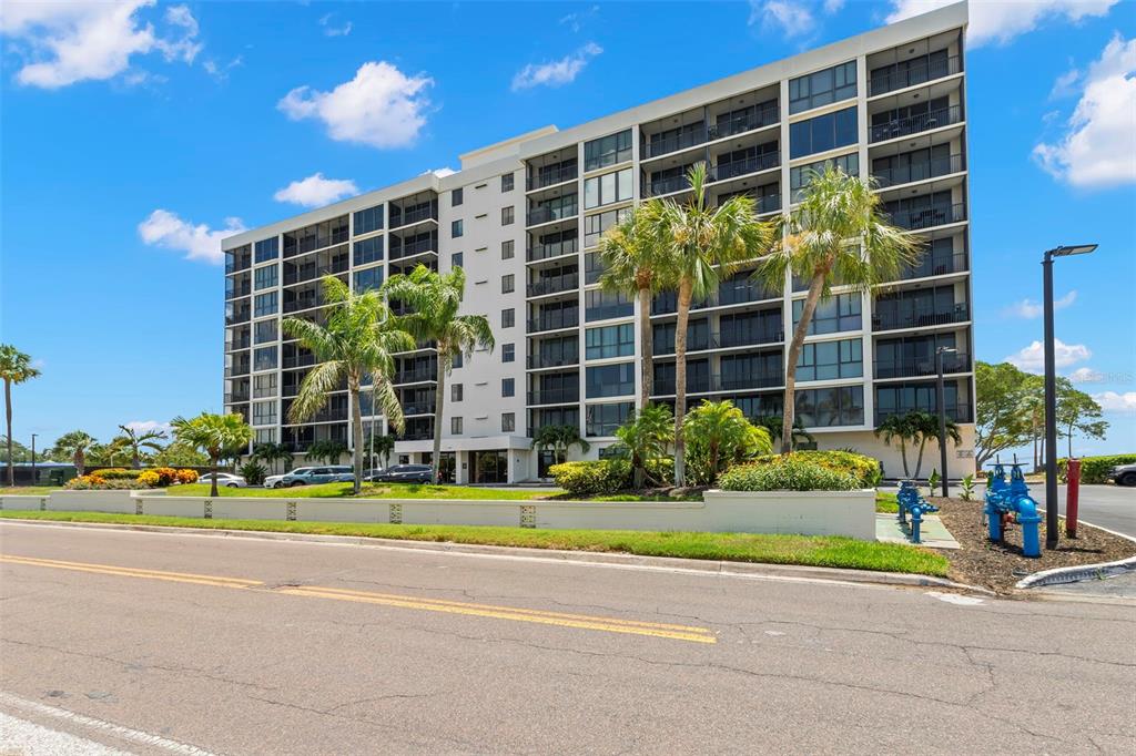 A multi-story apartment building with balconies stands behind palm trees and a paved road — 7100 SUNSHINE SKYWAY LANE S UNIT 703, ST PETERSBURG, FL