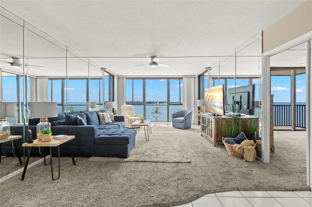 A living room with mirrored walls, a blue sectional sofa, and floor-to-ceiling windows overlooking water — 7100 SUNSHINE SKYWAY LANE S UNIT 703, ST PETERSBURG, FL