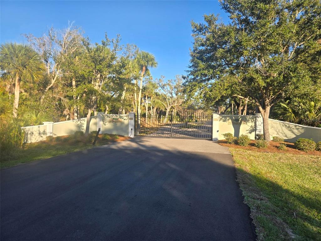 A paved driveway leads to a gated entrance flanked by stucco walls and mature trees — 9232 SPRING VALLEY ROAD, ENGLEWOOD, FL