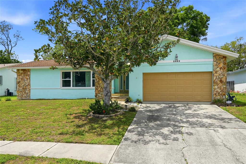 A light blue single-story house features a stone-accented facade, a two-car garage, and a tree — 2089 SWAN LANE, SAFETY HARBOR, FL