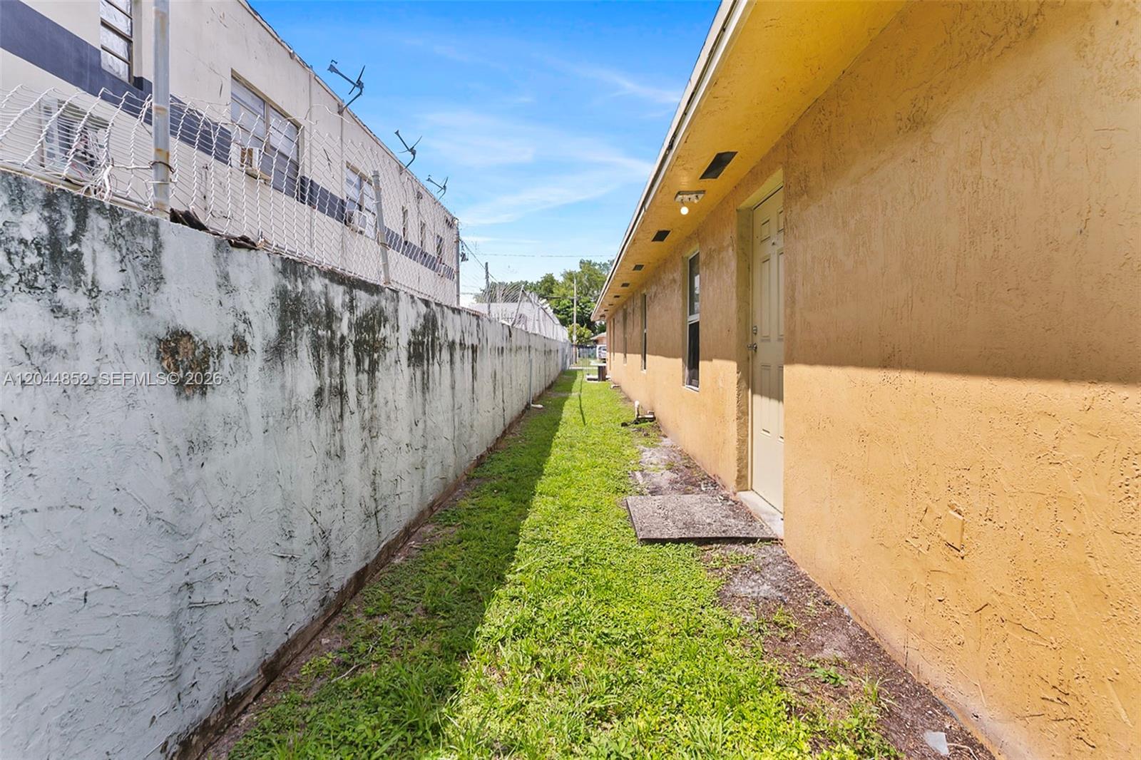 A narrow grassy alleyway sits between a stained concrete wall and a yellow building — 812 NW 70th St, Miami FL 33150, MIAMI, FL
