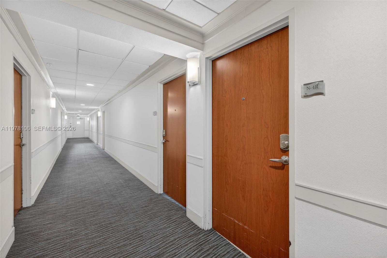 A long, carpeted hallway features white walls, drop ceilings, and several wooden apartment doors — 3801 S Ocean Dr # 4R, Hollywood FL 33019, HOLLYWOOD, FL