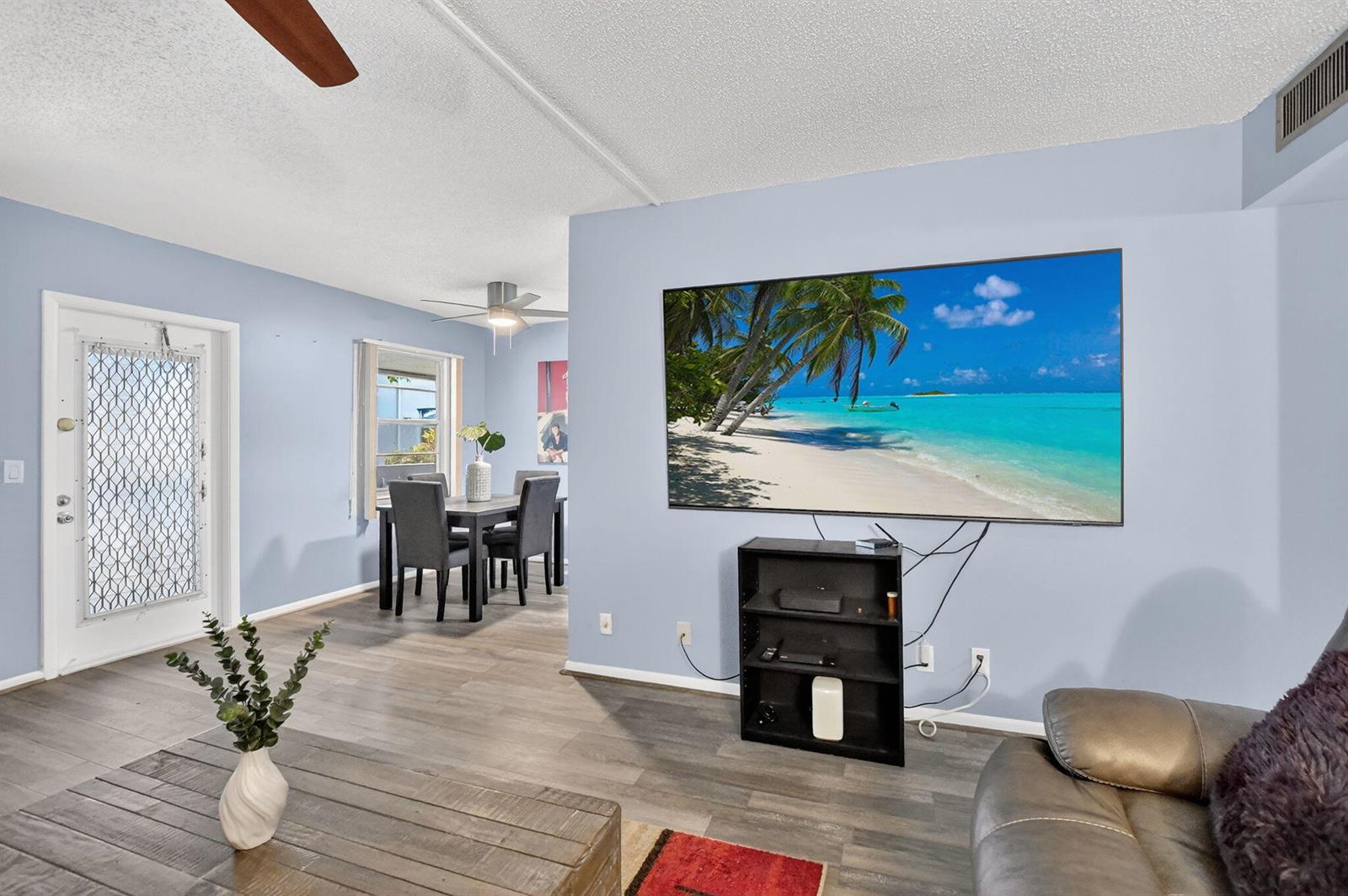 A living room with light blue walls, wood-look flooring, a wall-mounted television, and a dining area — 262 Ventnor R, Deerfield Beach FL 33442, DEERFIELD BEACH, FL