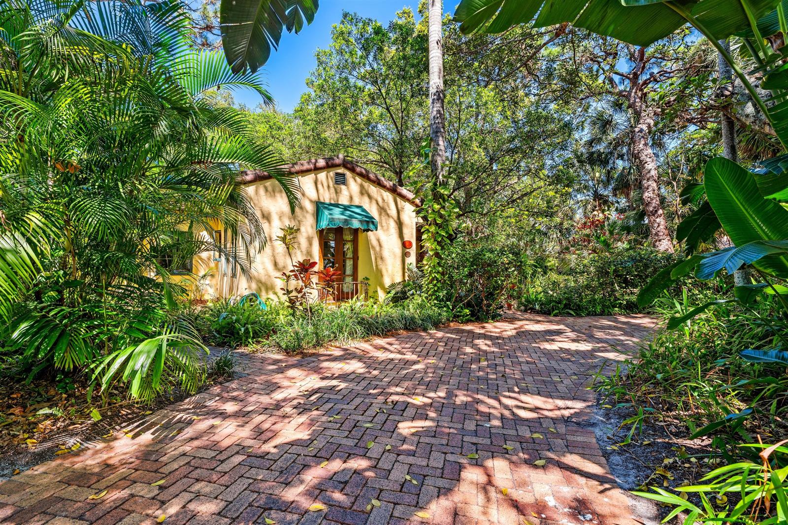 A yellow stucco cottage sits at the end of a brick driveway surrounded by lush foliage — 825 Alamanda Street, Boca Raton FL 33486, BOCA RATON, FL