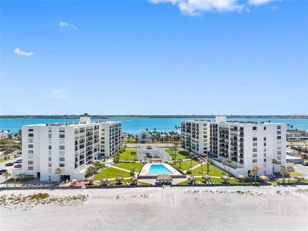 An aerial view shows two white apartment buildings flanking a central swimming pool and lawn — 1400 GULF BOULEVARD UNIT 101, CLEARWATER BEACH, FL