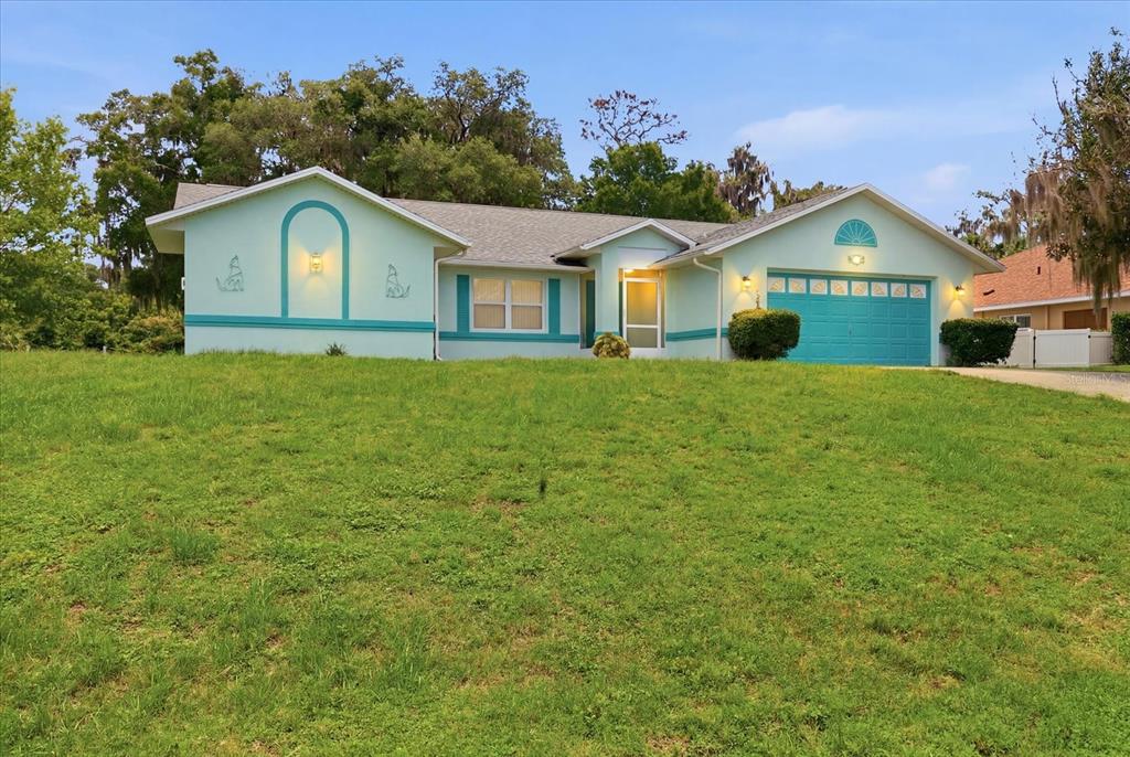 A light green single-story house with a teal garage door sits on a grassy hill — 1209 N TIMUCUAN TRAIL, INVERNESS, FL