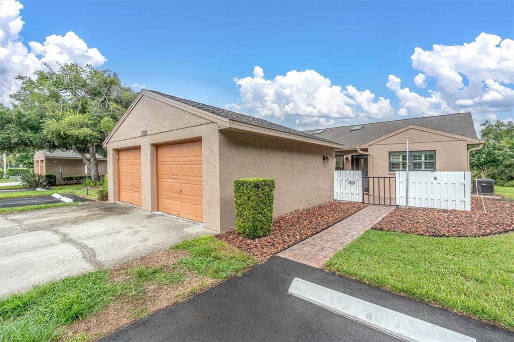 A tan single-story house features a two-car garage, a brick walkway, and a white fence — 3203 CHARTER CLUB DRIVE UNIT B, TARPON SPRINGS, FL