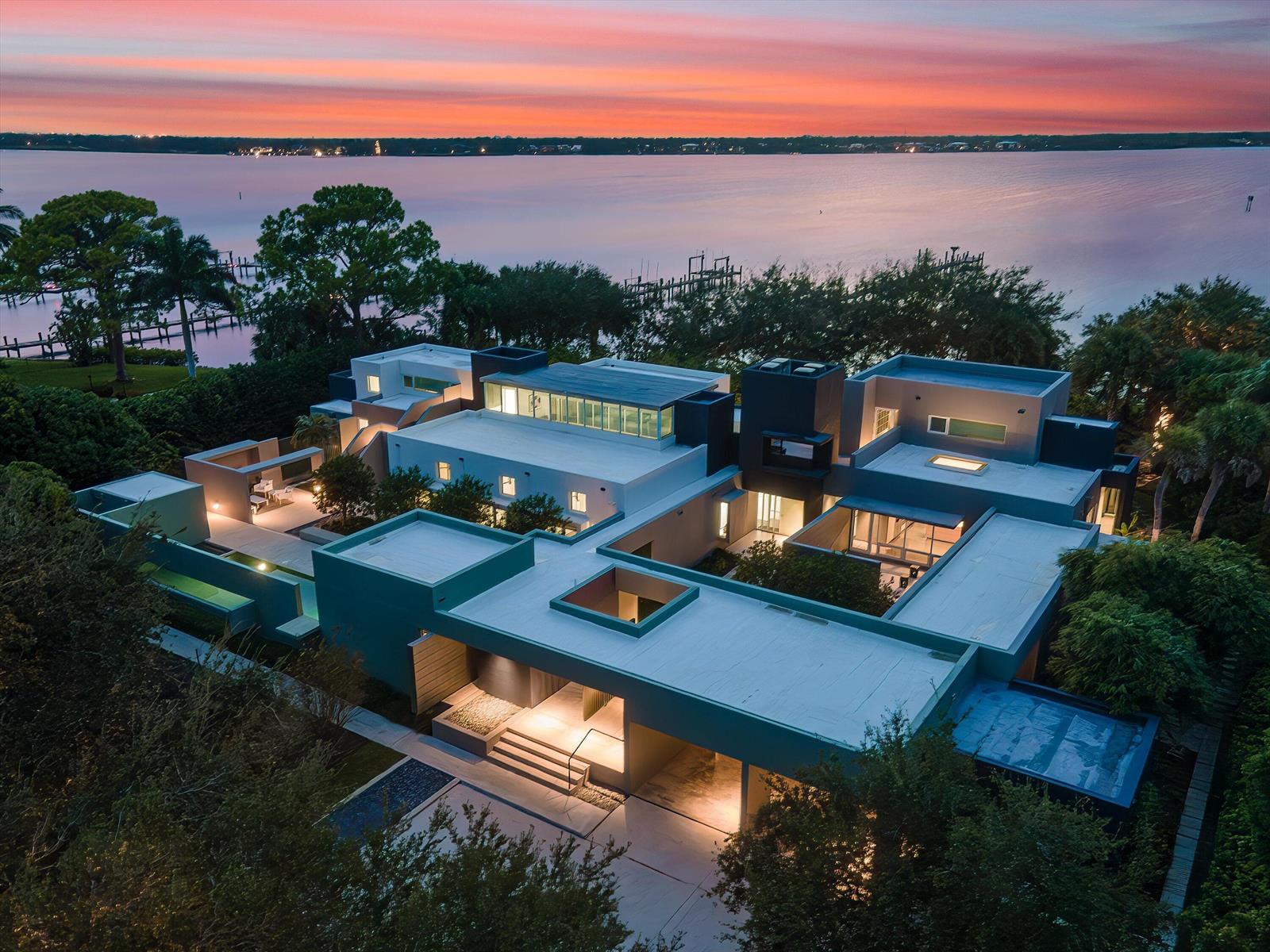 An aerial view of a modern flat-roofed mansion at sunset overlooking a large body of water — 1924 NW Shore Terrace, Stuart FL 34994, STUART, FL