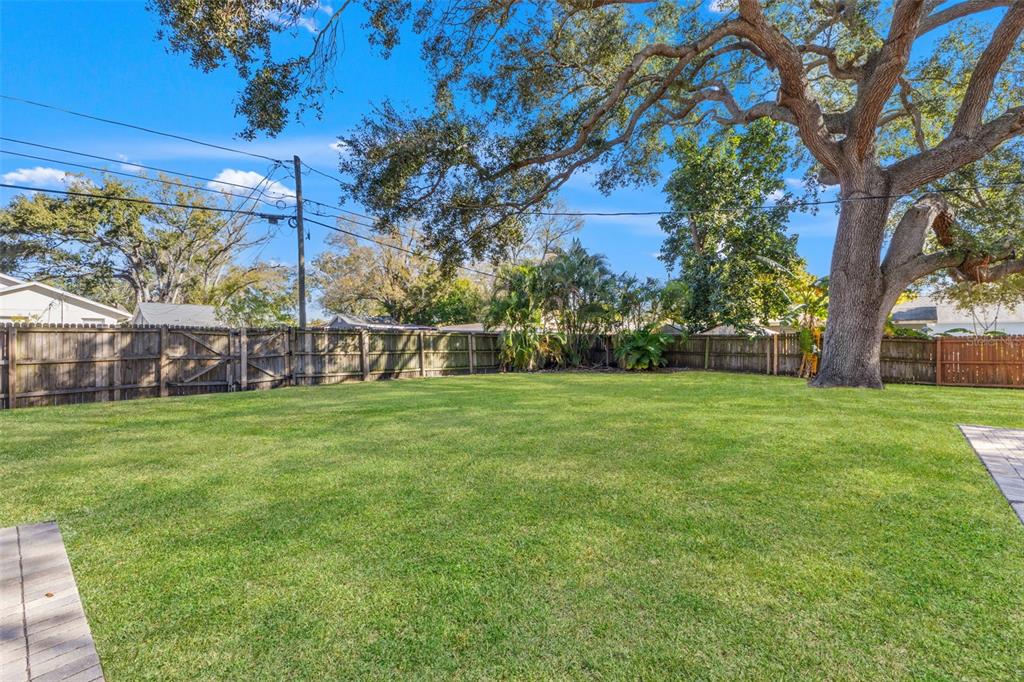 A large, grassy backyard features a wooden fence, mature trees, and stone patio borders — 1747 26TH AVENUE N, ST PETERSBURG, FL
