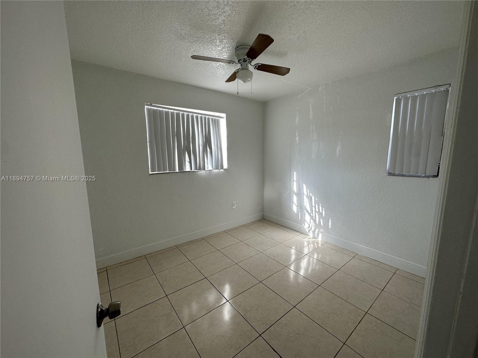 An empty room features white walls, light-colored tile flooring, a ceiling fan, and two windows — 1851 Ali Baba Ave # 4, Opa-Locka FL 33054, OPA-LOCKA, FL