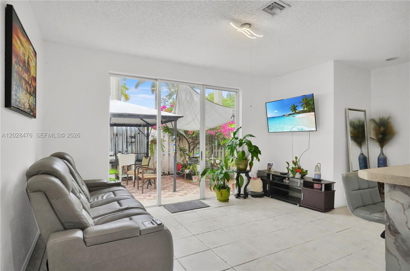 A living room features a grey sofa, tile flooring, a wall-mounted television, and sliding glass doors — HOMESTEAD, FL