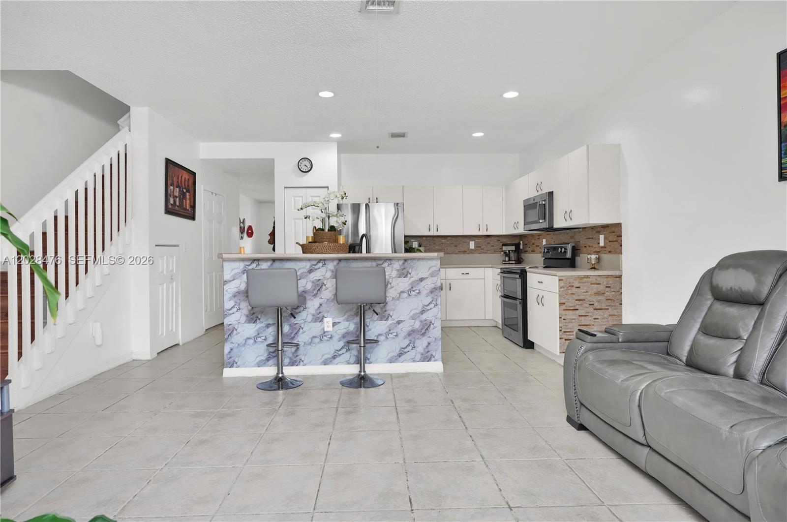 A kitchen with white cabinets and a marble-patterned island sits beside a gray sofa — HOMESTEAD, FL