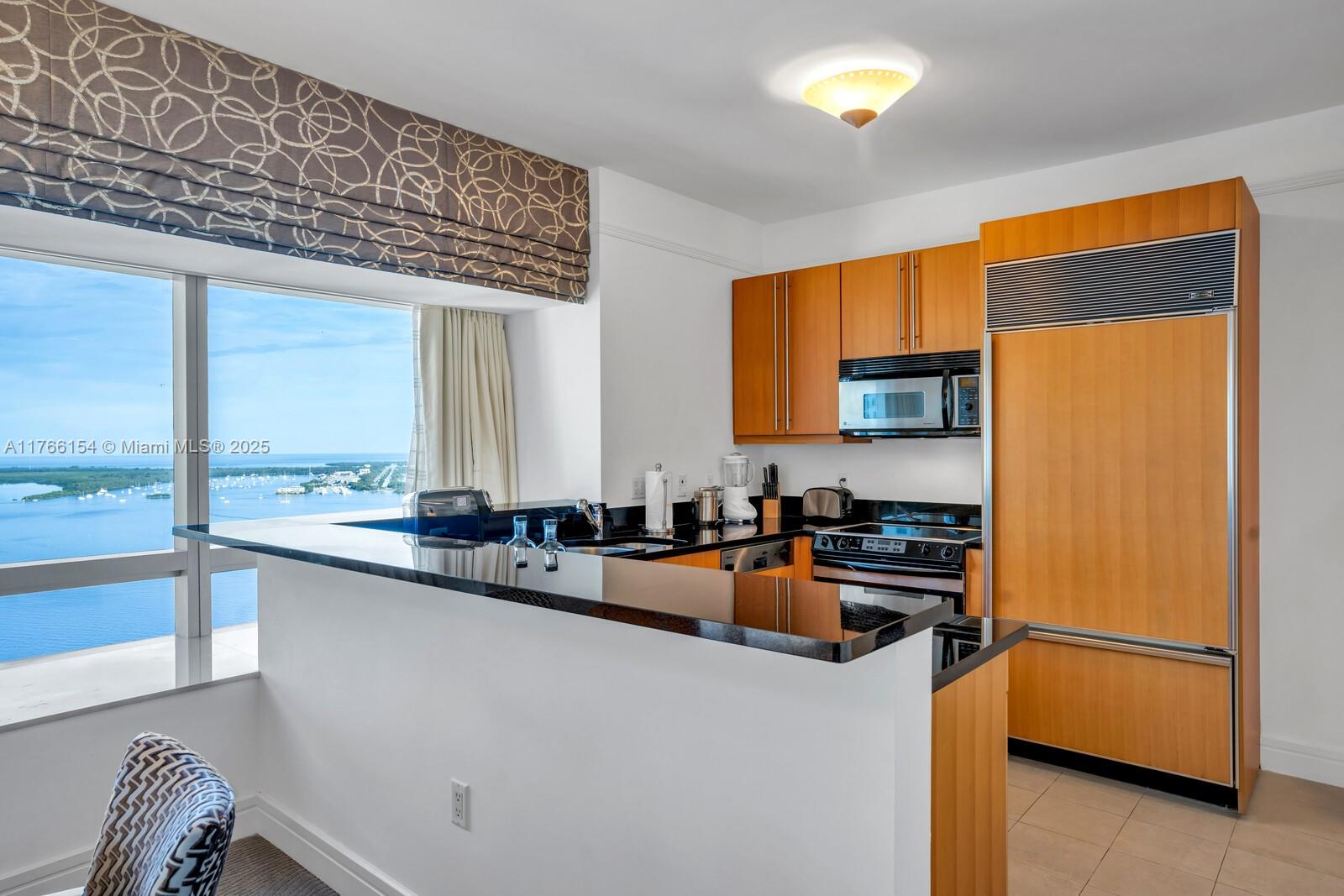 A kitchen with wood cabinets, black countertops, and a large window overlooking the water — 1435 Brickell Ave # 3607, Miami FL 33131, MIAMI, FL