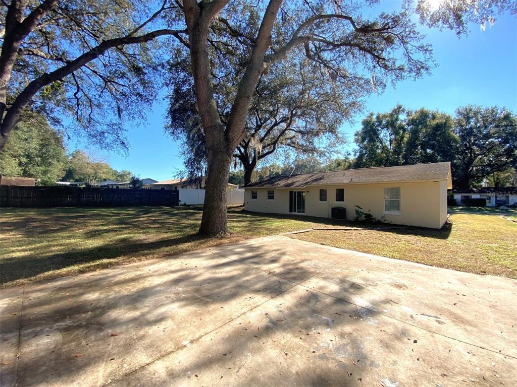 A concrete patio sits in the backyard of a single-story house with large oak trees — 11585 SE 129TH LANE, OCKLAWAHA, FL