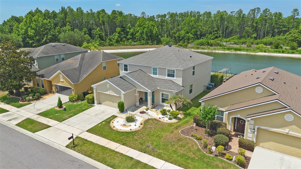 An aerial view shows three suburban houses with driveways and lawns bordering a small lake — 5521 SHASTA DAISY PLACE, LAND O LAKES, FL