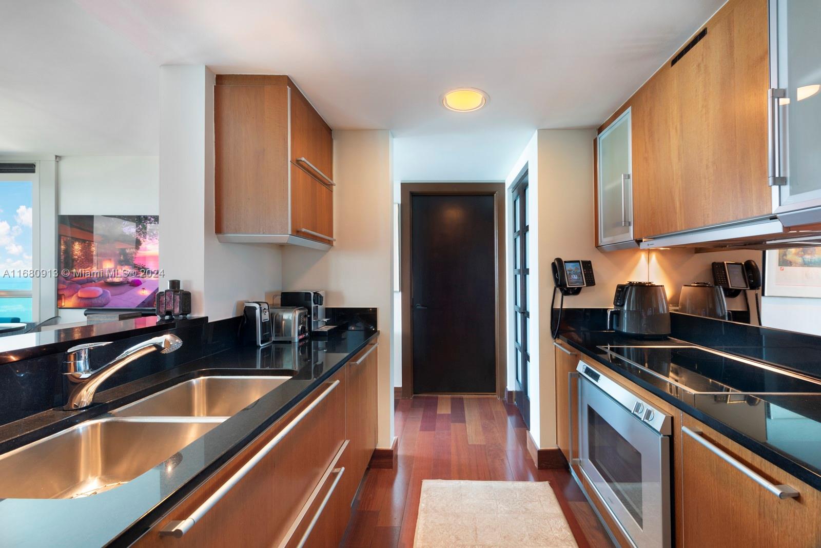 A galley kitchen features wood cabinetry, black countertops, stainless steel appliances, and hardwood flooring — 101 20TH ST # 2603, Miami Beach FL 33139, MIAMI BEACH, FL