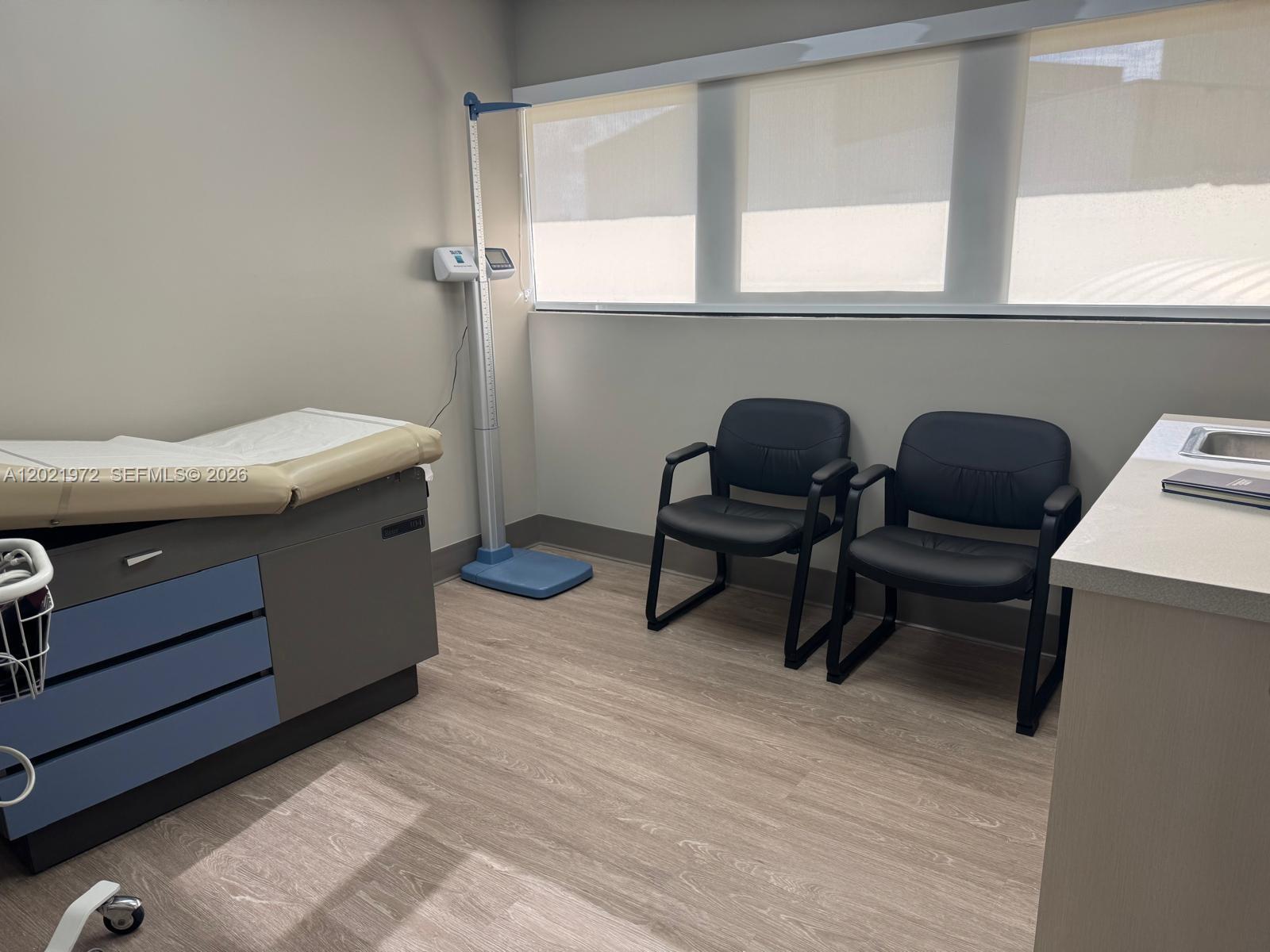 A medical examination room features a treatment table, a digital scale, and two black chairs — 7100 W 20th Ave # 214, Hialeah FL 33016, HIALEAH, FL