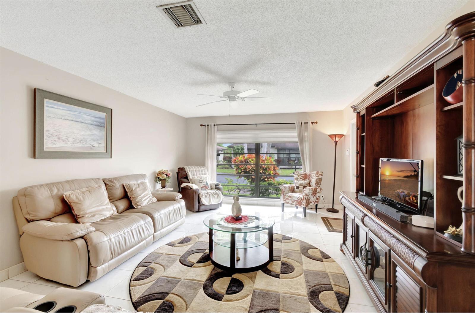 A living room features a beige leather sofa, a round patterned rug, and a wooden entertainment center — 4660 Greentree Place # 105b, Boynton Beach FL 33436, BOYNTON BEACH, FL