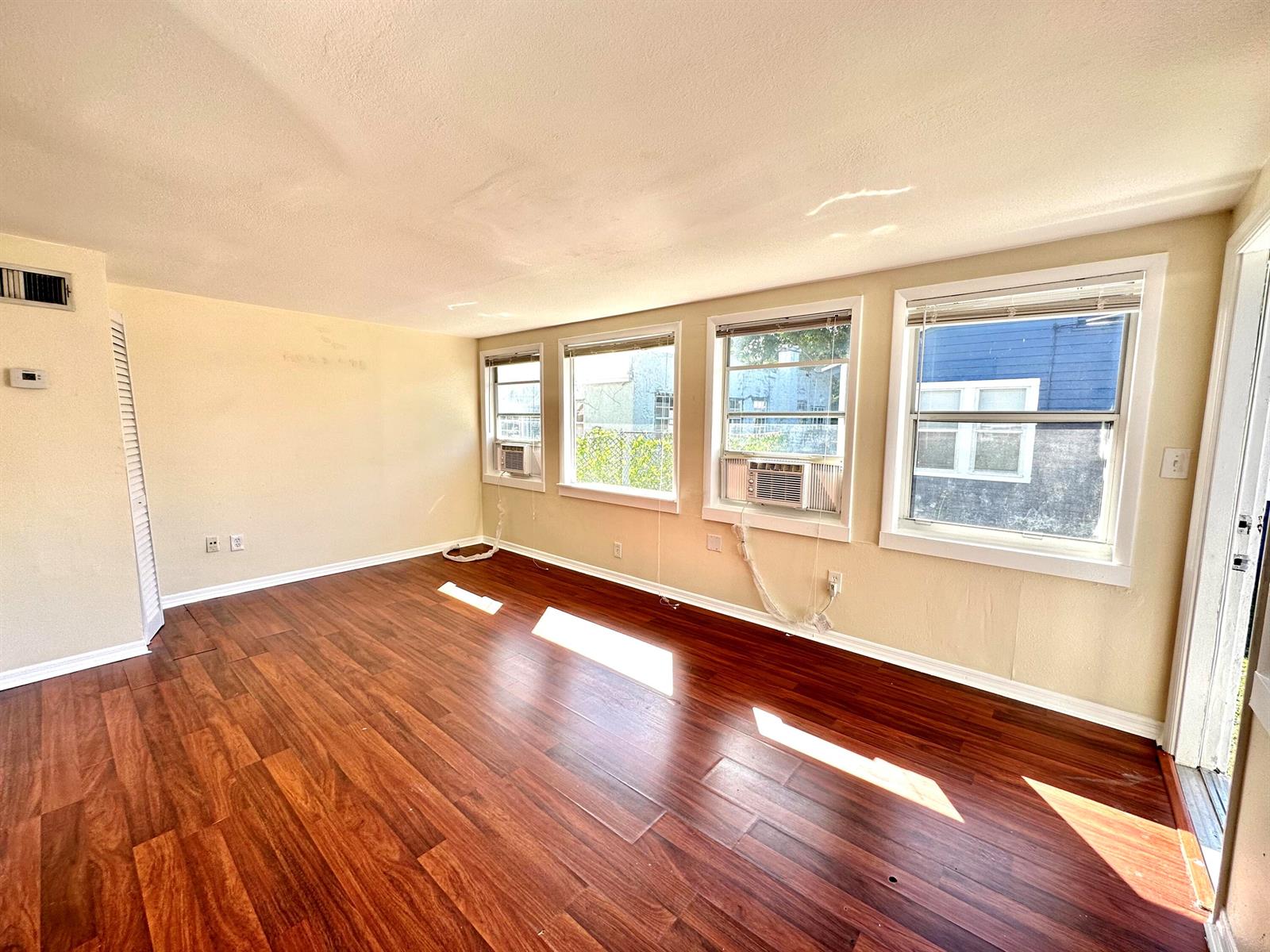 An empty room with dark wood-look flooring, beige walls, and three windows with air conditioners — 517 Kanuga Drive Unit 3, West Palm Beach FL 33401, WEST PALM BEACH, FL