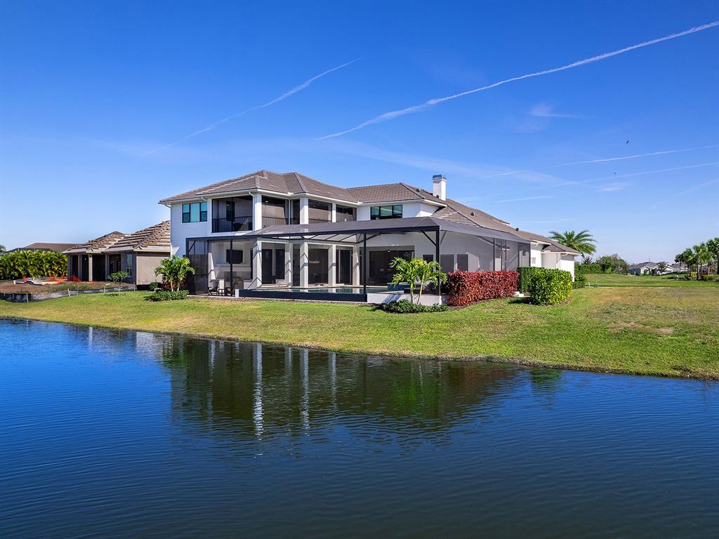 A two-story house with a screened-in pool area sits beside a lake under a blue sky — 4218 BUTTE TRAIL, LAKEWOOD RANCH, FL